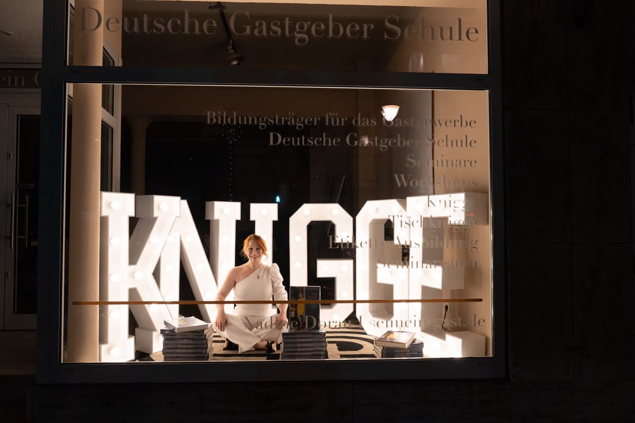 Das leuchtende KNIGGE-Schaufenster der Deutschen Gastgeber Schule bei Nacht, Nadine sitzt zwischen den Buchstaben